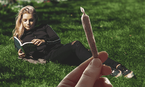 ruka drziaca konopnu cigaretu a zena ktora lezi na trave a cita knihu