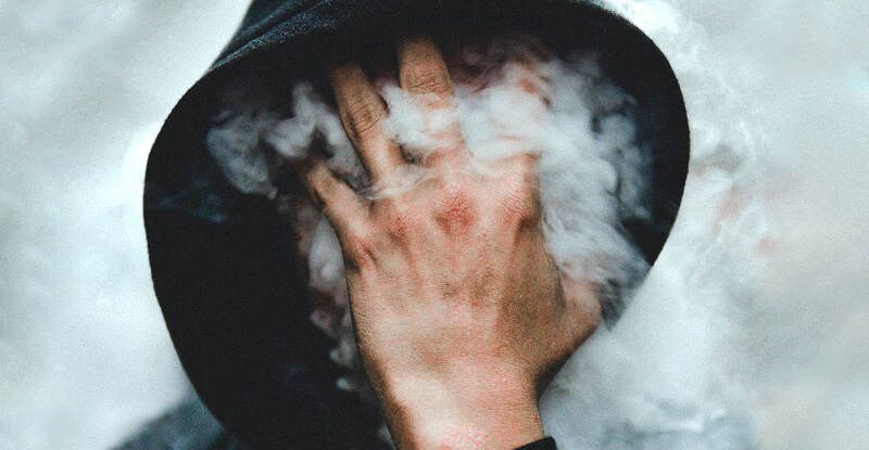Človek v kapucni si skrýva tvár dlaňou, spoza ktorej uniká množstvo dymu
