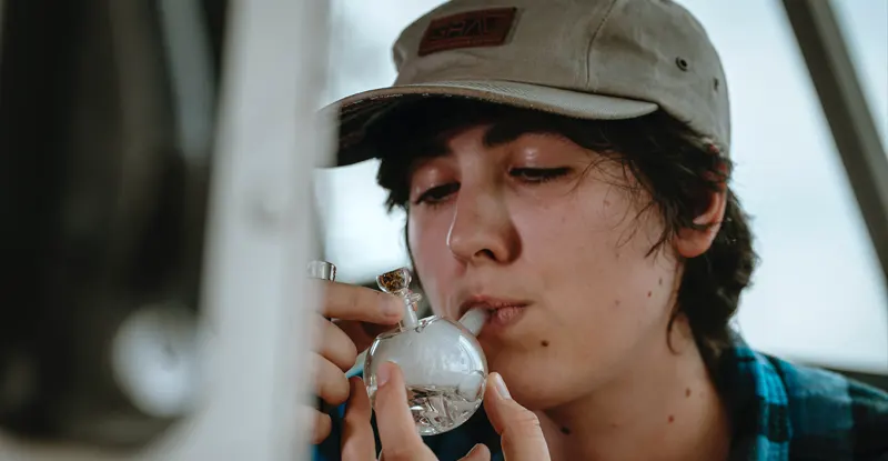 mladý človek so šiltovkou na hlave fajčí marihuanu z malého skleneného bonga