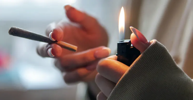 Detail ženských rúk - v jednej ruke ubalená jointová cigareta a v druhej horiaci zapaľovač