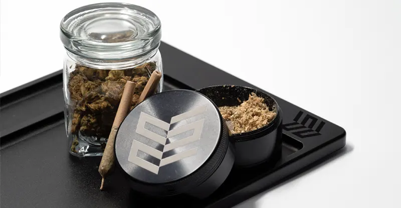 Čierny podnos, sklenený pohár so šiškami marihuany, jointy, drvička a krabička s podrvenou bylinou