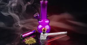 Sklenené bongo fialovej farby, fialový kovový clipper zapaľovač, sklenená fajka s hlavičkou v tvare lebky a nadrvená marihuana