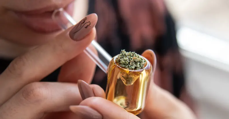 Detail sklenenej fajky s kotlíkom plným podrvenej marihuany, ktorú si prikladá k ústam žena s upravenými nehtami
